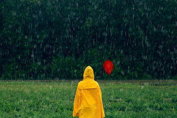 Boy in yellow raincoat with red balloon standing in the middle during rainfall in front of coniferous forest on rainy, foggy day. Travel lifestyle concept vacations outdoor. Horror movie concept.