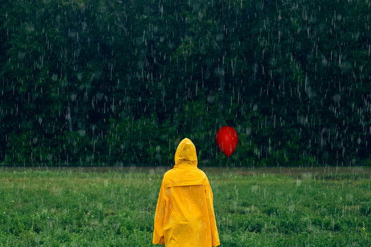Boy in yellow raincoat with red balloon standing in the middle during rainfall in front of coniferous forest on rainy, foggy day. Travel lifestyle concept vacations outdoor. Horror movie concept.