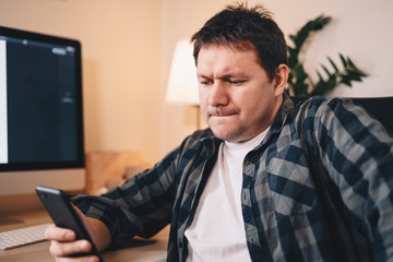 Freelancing young man typing on his mobile phone, being in a stresseful situation at home office.