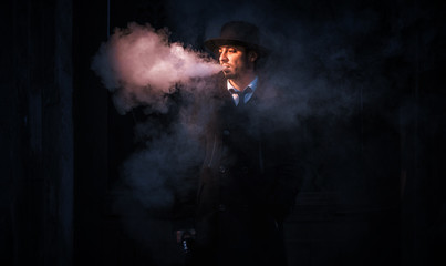 Young hipster man smokes or vaping an electronic cigarette. Man wearing a tie and black hat. Delicious pairs. Cloud of steam.