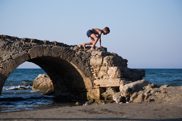 Fototapeta premium Little boy plays on a ruined old stone bridge next to a beach.