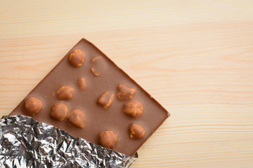 Bar of chocolate with hazelnuts and covered with foil lays on light wooden background.