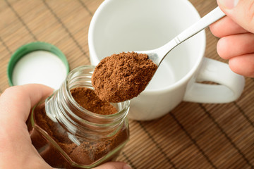 White cup; jar of coffee and a green cover lays on brown background. Taking some coffee from a jar and putting it into the cup. Making coffee