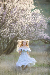 Obraz na płótnie Canvas A young long-haired blonde is walking in a blooming garden. Dress and hair fluttering like in a fairy tale