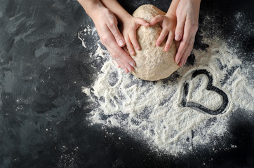 Mother and child hands prepares the dough with flour on dark wooden table. Bakery background. Heart...