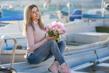 Beautiful girl in spring or autumn, by the sea in the boat Bay. With a bouquet of flowers in his hands and tight stylish jeans and a pink sweater.