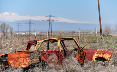 Abandoned and rusty wreckage of an yellow vintage Soviet Russian car in the middle of dry hay with scenic ice top mountains and clear blue sky on the background in rural Southern Armenia.