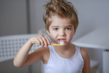 Close up portrait of cute, adorable, toddler boy wearing denim overalls, long T-shirt, sitting on the floor, brushing his teeth with a toothbrush