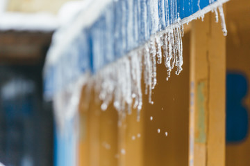 Cropped photo of icicles hanging from roof of the building, drops are dripping from them/ danger for humans, sudden warming, poor thermal insulation, ice stalactite, formation of icicles.