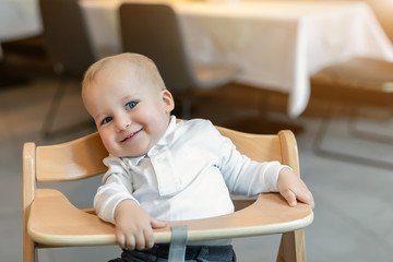 Cute little baby boy in white polo t-shirt sitting in wooden baby chair and laughing at cafe indoors. Portrait of adorable blond kid having fun at restaurant