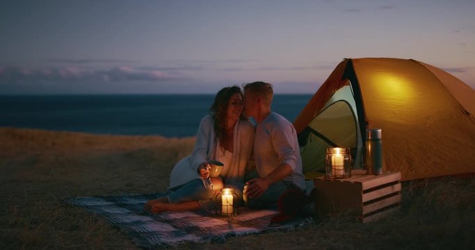 Attractive couple snuggling with candles and tent in beautiful fields at dusk, camping romance, adventure