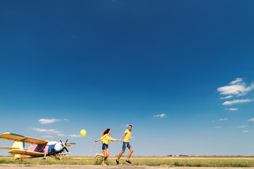Obraz na płótnie Canvas Cute couple running and holding hands. In background field and airplane. Dominating blue and yellow colors.