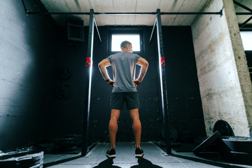 Muscular man in sportswear preparing to do chin-ups while standing with hands on hips in cross-fit gym. Back turned.