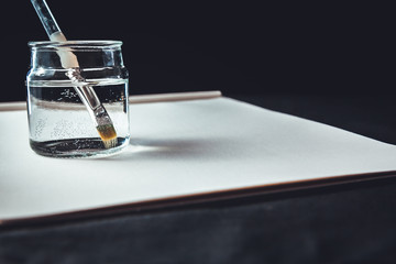 Blue Paintbrush With Gold Bristles Resting In A Glass Jar of Water Next To A Blank Canvas