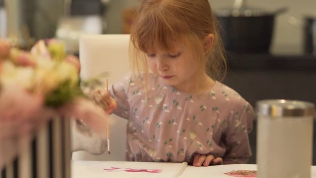 A close up portrait video of little girl drawing a pink flower in the notebook. She uses watercolours and a brush. The background is blurred.
