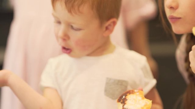 A little redhead boy eats a big cookie. The powdered sugar is on both of his lips. He looks somewhere in the right direction. His mother checks on him from time to time. It is close up portrait video