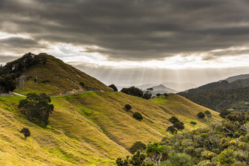 The rays of the sun pierce through the gap between the gray clouds, illuminating the hilly...