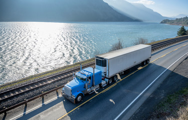 Blue classic American bonnet big rig semi truck trancporting cargo in refrigerated semi trailer moving on the road along the river in Columbia River Gorge area © vit