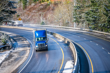 Blue day cab big rig semi truck with semi trailer transporting goods on winding wet winter snow road