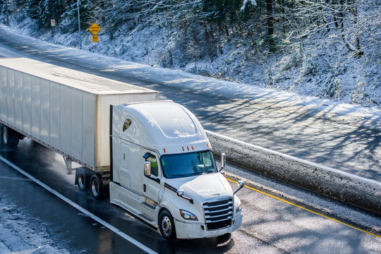 Bright white modern big rig semi truck transporting dry van semi trailer running on winter snowy wet road