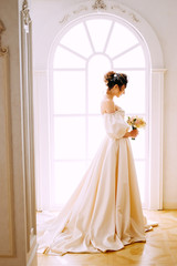 Elegant young woman in creamy satin dress standing near the window in the chapel before getting married