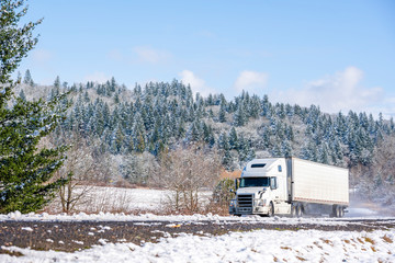 White big rig long haul semi truck with refrigerator semi trailer running on the winding winter snowy road with forest