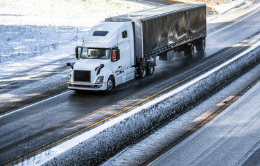 White big rig semi truck with frame covered black semi trailer running on the wet winter snow...