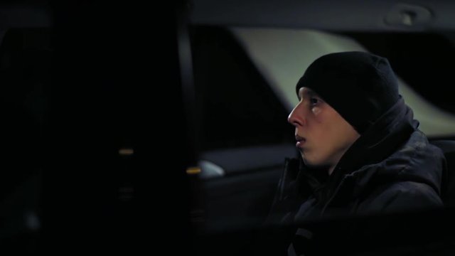 Profile of young man wearing black hat and jacket shaking his head while sitting in the car at night.