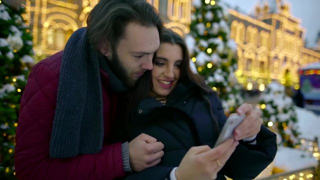 young lovers are watching their selfies in mobile phone, standing on square with new year decoration