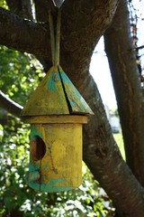 A brightly painted bamboo birdhouse in colors of green and yellow hangs from the branch of a leafy tree.