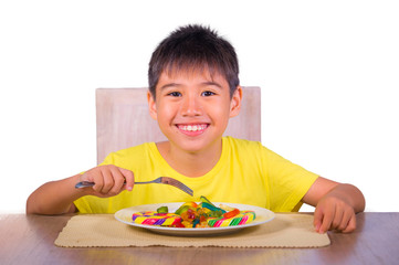 young happy and excited male kid smiling cheerful eating dish full of candy and lollipop sitting at table isolated on white background in sugar addiction and abuse