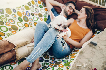 Obraz premium happy hipster couple with bulldog relaxing in hammock on the beach in sunset light, summer vacation. stylish family with dog cuddling and having fun, cute moments in summer evening