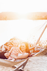 Young family with baby on the beach, smiling father and mother holding cute little girl while lying in a hammock, hipster family on vacation near lake at sunset