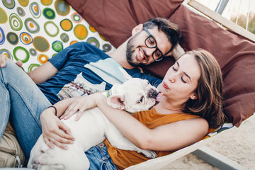Cute couple lying in a hammock with a dog, funny white bulldog relaxing with a hipster young family...
