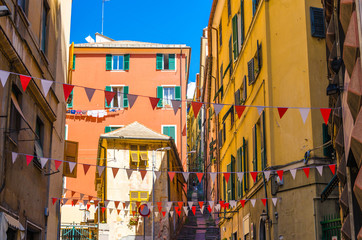 Fototapeta na wymiar Small red and white flags hang above street with colorful multicolored buildings with shutters on windows in historical centre of old european city Genoa (Genova), Liguria, Italy
