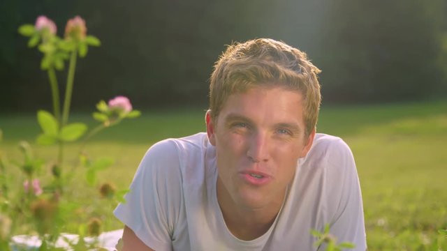LENS FLARE, DOF, PORTRAIT: Happy boyfriend blows a kiss at the camera after filming a video for girlfriend while lying in the idyllic grassy field. Cheerful digital nomad filming a v-log in nature.