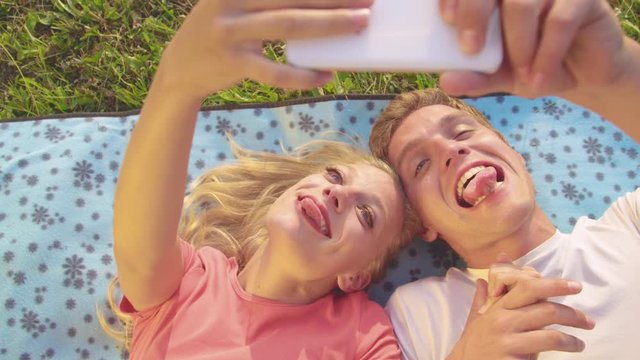 SLOW MOTION, CLOSE UP: Excited young woman and handsome man making funny faces and taking selfies while lying on a blanket in the middle of a meadow on a warm summer day. Goofy couple taking photos.