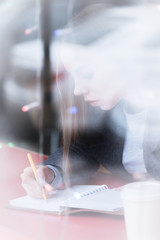 Portrait of an attractive young journalist girl with pen and notepad in cafe behind the showcase. Non-contrast view through window glass reflection