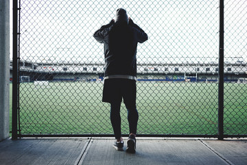 Tired sportsman leaned and looking throurh the rabitz fence on football field. Runner resting near the fence