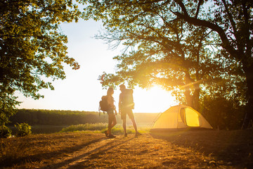 Fototapeta premium A man and a woman in a hiking trip with backpacks near a tent at sunset. Honeymoon in nature