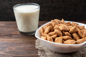 Dried breakfast pads in white bowl and milk on dark wooden background.