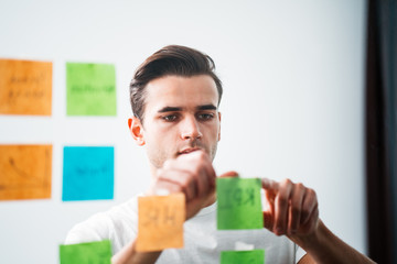 Concentrated businessman standing at office space behind glass wall with sticky notes. Creative professional manager create new business ideas