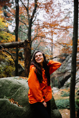 Young woman wearing warm orange sweater in the autumn forest. Misty landscape with mossy rocks. Cute smiley woman in the nature. Autumn forest hiking  - 248285396