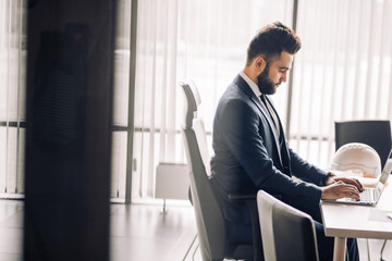 Photo of caucasian brunette bearded man 30s in formal suit sitting alone in well-lit office and working on laptop against the window