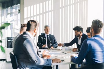 Indian business adviser analyzing company financial report shows documents with charts to Senior grey-haired investor discussing balance sheet data during corporate meeting