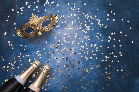 Two champagne bottles, golden carnival mask and confetti stars  on blue background. Christmas background, top view, copy space.