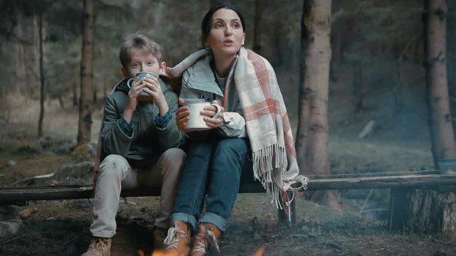 Mother and son sitting by campfire with mugs of hot beverage talking