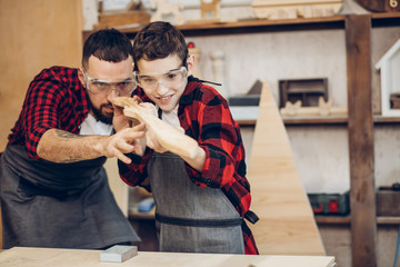 Fatherhood, hobby, carpentry, woodwork and people concept - Father and little son wearing protective glasses testing hand-made wood sword smooth surface at workshop