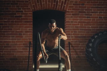 African muscular shirtless man is concentrated on the exercise with battle rope requiring increased...