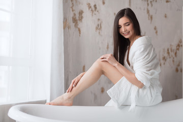 Brunette spa caucasian girl with white young skin, wearing white bathing robe, preparing to bath, looking down, enjoying warm relaxing water over light background.